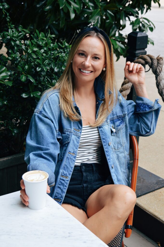 Woman with blonde hair sitting at a small table holding a paper cup of coffee. Woman is wearing a denim jacket and black headband.